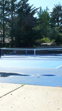 Yakın zamanda temizlenmiş bir picleball sahasından su buharlaşması güneşli bir günde ağaçlarla çevrili ve arka planda bazı oyuncularla ilginç desenler oluşturur.