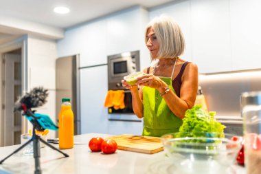 Önlük giymiş yaşlı bir kadın modern mutfağında sağlıklı yiyecekler hazırlıyor ve akıllı telefonuyla video tarifi kaydediyor.