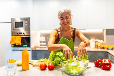 Önlük giymiş yaşlı bir kadın. Parlak bir mutfakta taze malzemelerle salata yapıyor. Sağlıklı yaşam tarzını gösteriyor ve evde yemek pişiriyor.