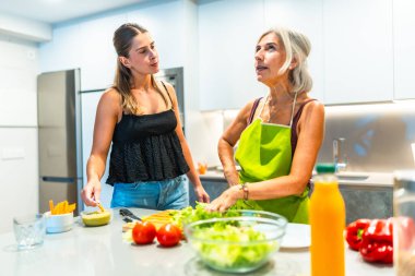 Modern bir mutfakta yemek hazırlayan iki kadın taze sebzelere ve sağlıklı yeme alışkanlıklarına odaklanıyor ve aile bağlarını güçlendiriyor.