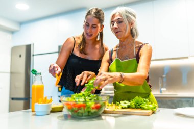Anne ve kız modern bir mutfakta marul, domates ve portakal suyuyla karışık bir salata hazırlıyorlar. Birlikte vakit geçiriyorlar ve sağlıklı beslenme alışkanlıklarını destekliyorlar.
