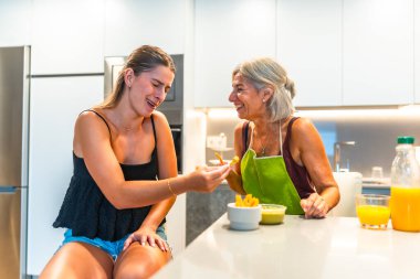 Büyükanne ve torun birlikte gülerken modern bir mutfakta patates kızartması ve guacamole yiyorlar.