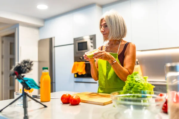 Önlük giymiş yaşlı bir kadın modern mutfağında sağlıklı yiyecekler hazırlıyor ve akıllı telefonuyla video tarifi kaydediyor.