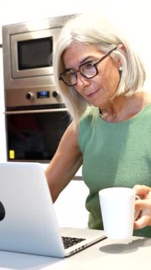 Uzaktan kumandayla çalışan, bilgisayarla yazı yazan ve modern mutfakta kahvenin tadını çıkaran olgun bir iş kadını.