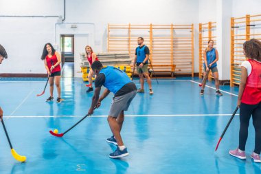 Spor salonunda antrenman yaparken paslaşan takım oyuncuları, canlı mavi zeminde sopalarıyla ustaca topa vuruyorlar, takım çalışması ve atletizm sergiliyorlar.