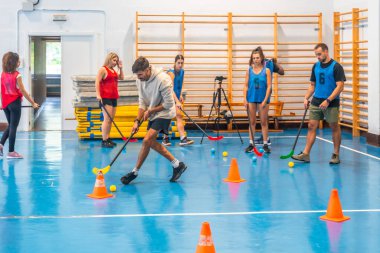 Bir grup sporcu florbol antrenmanı yapıyor, sopayla pas veriyor ve spor salonundaki konilerin etrafında manevra yapıyor, takım çalışması ve atletizm sergiliyor.