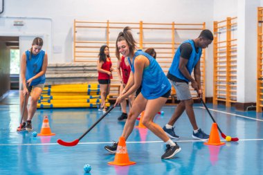 Mavi tişört giyen kadın ve erkek sporcular, mavi zemin ve sarı ekipmanlı bir spor salonunda turuncu koni etrafında sopalarla top sürüyor.
