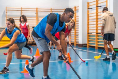 Floorball oyuncuları, iyi ışıklandırılmış bir jimnastik salonunda sopa tutma becerilerini geliştirirken, takım çalışması ve atletizm sergiliyorlar.