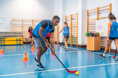 Spor salonunda top sürmeye odaklanmış genç bir sporcu antrenman sırasında takım arkadaşlarıyla sopa tutma becerileri, çeviklik ve hassasiyet gösterisi.