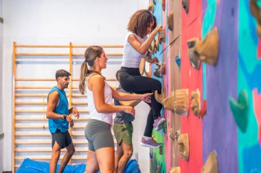 Genç bir kadın spor yaparken spor yapıyor spor salonundaki renkli bir duvara tırmanıyor. Eğitmeni onu desteklerken diğer sporcular arka planda antrenman yapıyor.
