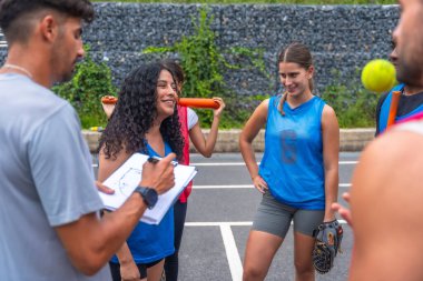 Koç, açık hava sahasında antrenman yaparken gülümseyen bayan beyzbol takımına oyun stratejisini, takım çalışmasını, sportmenliği ve atletizmi anlatıyor.