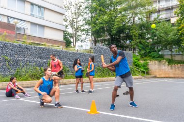 Şehir ortamında beyzbol oynayan bir grup genç arkadaş, takım arkadaşları sıralarını beklerken bir erkek oyuncu topa vuruyor.