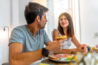 Genç bir çift mutfak masasında yemek yiyip sohbet ediyor. Birlikte rahatlatıcı bir sabah geçiriyorlar.