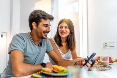 Genç çift gülümsüyor ve rahat mutfaklarında kahvaltıdan zevk alıyor, akıllı bir telefonla bağlanıyor ve anlarını online olarak paylaşıyorlar.