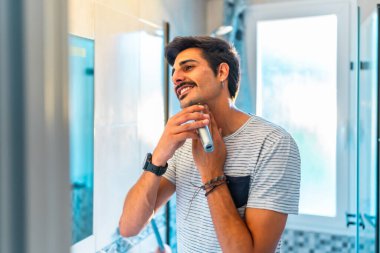 Banyodaki aynada sakalını kesen genç adam elektrikli jilet kullanıyor ve sabah rutininin tadını çıkarıyor.