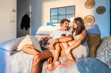 Genç bir çift evdeki kanepede dinleniyor, dizüstü bilgisayarda gezinirken günbatımının sıcak ışığının pencereden içeri girmesinin keyfini çıkarıyorlar.