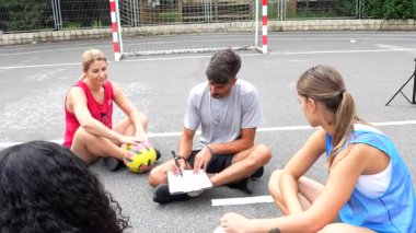 Futbol koçu stratejik rehberlik ve taktiksel içgörü sağlıyor, oyun diyagramları çiziyor ve güneşli alan antrenmanında bayan sporcuları motive ediyor.