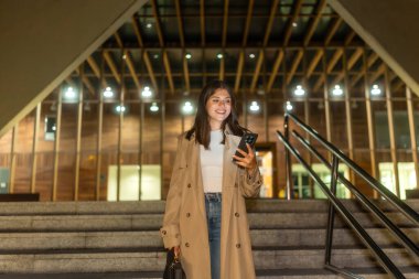 Genç bir kadın akıllı telefonunu kontrol ederken gülümsüyor, gece şehir merdivenlerinden iniyor, trençkot giyiyor, modern iletişimi ve şehir hayatını yansıtıyor.