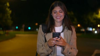 Geceleri şehirde gezen neşeli genç bir kadın. Akıllı telefonuyla mesajlaşırken gülümsüyor. Sonra da şehir ışıkları ve arka planda trafik olan bir telefona mutlu bir şekilde cevap veriyor.