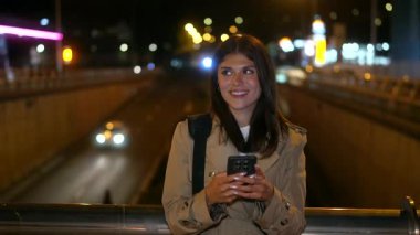 Cep telefonuyla mesajlaşırken gülümseyen neşeli genç bir kadın, şehir köprüsünde dururken trafik ışıkları ve araba farları arka planda güzel bir bokeh etkisi yaratıyor.