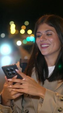 Neşeli genç bir kadın gülümsüyor ve şehirde akıllı telefonuyla fotoğraf çekiyor. Bulanık renkli sokak ışıklarının arka planında mutlu bir anı yakalıyor.