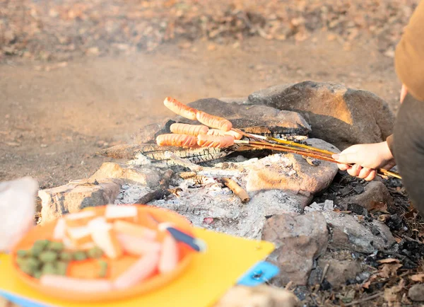 Sausages are fried over a small fire. Grilling sausages over a campfire in the forest. Picnic with family in the countryside concept.