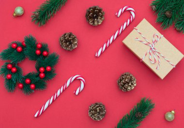 Kırmızı arka planda iki şeker kamışı, böğürtlenli Noel çelengi, hediye kutusu, köknar konileri ve Noel süslemeleri. Yeni yıl tebrik kartı. Noel, yeni yıl konsepti. Düzleştirme biçimi.