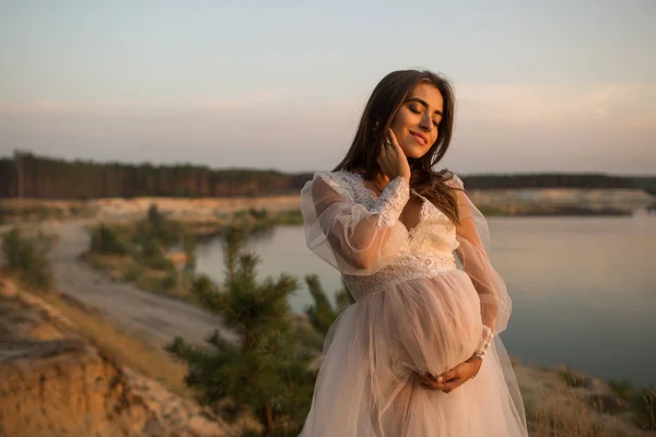 Pregnant girl posing near the river at sunset. Young pregnant woman holds her hands on her swollen belly. Love concept.