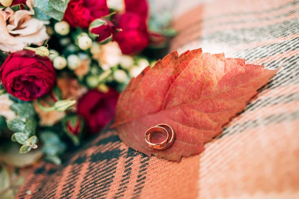 Wedding rings on autumn leaf. Accessories for the autumn wedding. Beautiful wedding bouquet of red and beige roses.