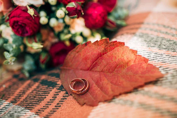 Wedding rings on autumn leaf. Accessories for the autumn wedding. Beautiful wedding bouquet of red and beige roses.