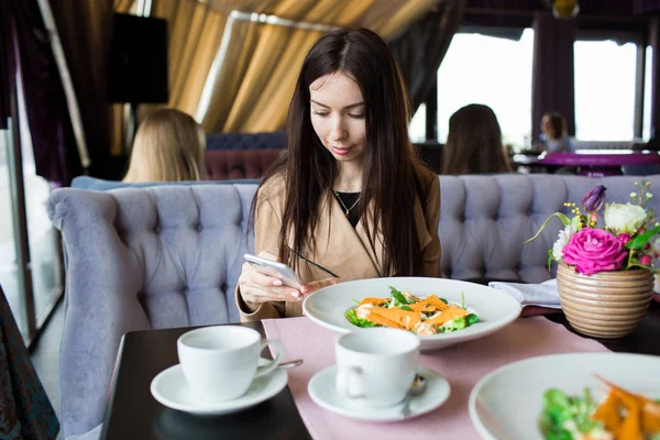 Hermosa chica en un café con un teléfono en sus manos. Primer plano, sentada en un café-restaurante, esperando su orden . — Foto de Stock