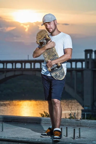 Güzel bir gün batımı arka plan üzerinde kaykay ile ayakta genç şık kaykaycı. Aktif yaşam tarzı.