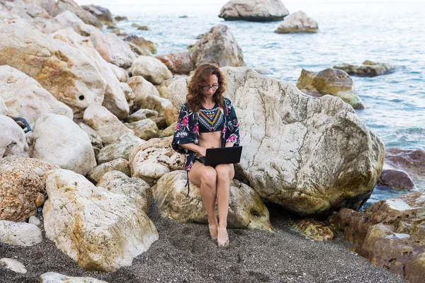 Woman workaholic works on netbook sitting on a stone on the beach.