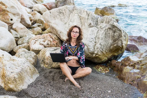 Mujer adicta al trabajo trabaja en netbook sentado en una piedra en la playa . — Foto de Stock