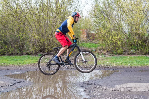 Un cycliste en short rouge et une veste jaune à vélo sur la roue arrière à travers une flaque d'eau . — Photo