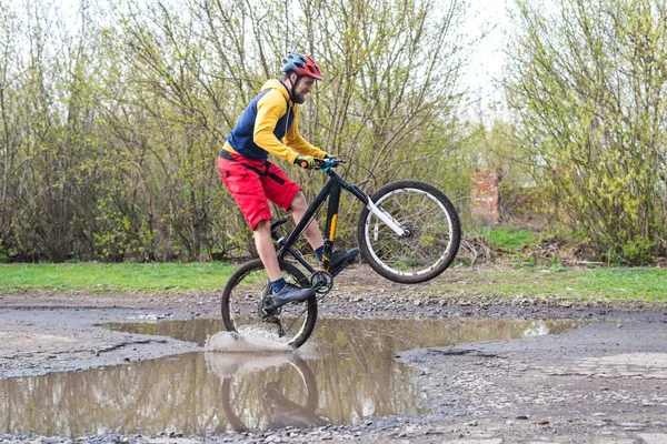 Un cycliste en short rouge et une veste jaune à vélo sur la roue arrière à travers une flaque d'eau . — Photo