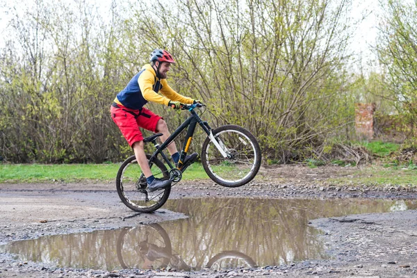 Un cycliste en short rouge et une veste jaune à vélo sur la roue arrière à travers une flaque d'eau . — Photo