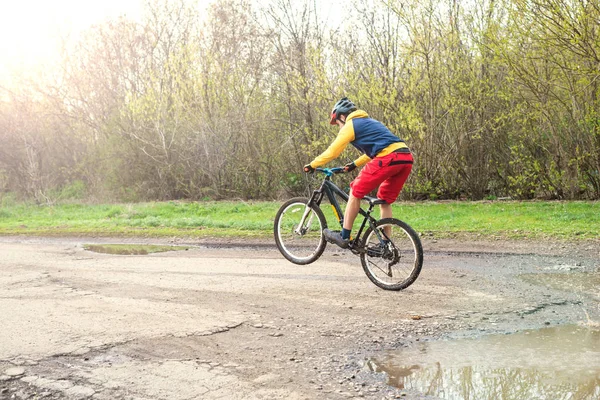 Un ciclista en pantalones cortos rojos y una chaqueta amarilla montando una bicicleta en la rueda trasera a través de un charco . — Foto de Stock