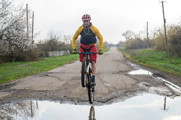 Un cycliste en short rouge et une veste jaune à vélo le long d'une rue avec des flaques d'eau . — Photo