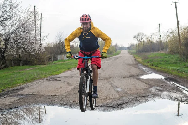 Un cycliste en short rouge et une veste jaune à vélo sur la roue arrière à travers une flaque d'eau . — Photo