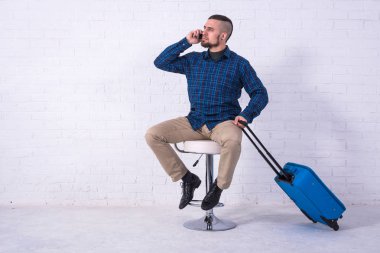 Mavi bavullu bir adam beyaz tuğla duvarın yanındaki sandalyede oturuyor ve telefonda konuşuyor..