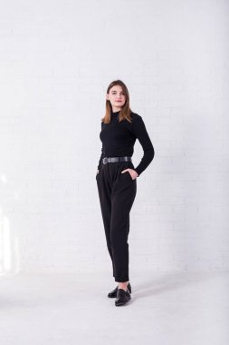 Siyah kazaklı, pantolonlu, beyaz tuğlalı duvarın yanında duran esmer bir kız..