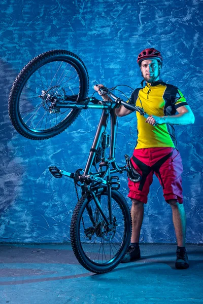 Cyclist in a yellow T-shirt and red shorts is holding a mountain bike against the background of a blue wall.