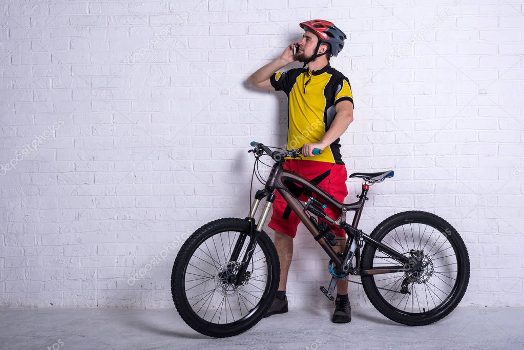 Cyclist in a yellow T-shirt and red shorts sitting next to a mountain bike and talking on a smartphone against the background of a white wall, free space.