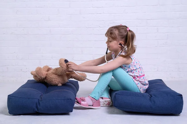 Little girl playing doctor and listening teddy bear with stethoscope against a white brick wall.