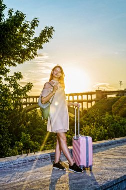 Genç güzel kız pembe bir arka plan üzerinde bir bavul ile duran güzel bir gün batımı.