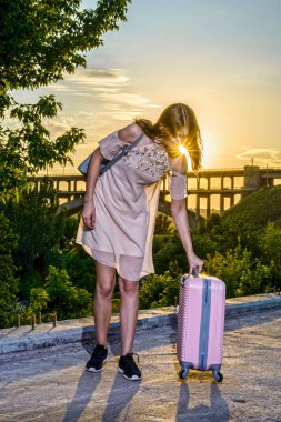 Genç güzel kız pembe bir arka plan üzerinde bir bavul ile duran güzel bir gün batımı.