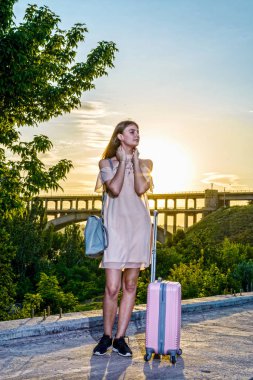Genç güzel kız pembe bir arka plan üzerinde bir bavul ile duran güzel bir gün batımı.