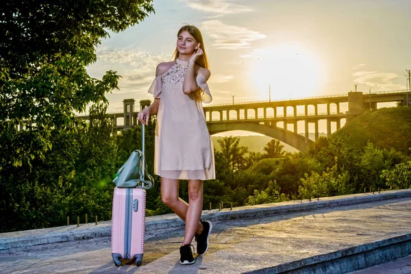 Genç güzel kız pembe bir arka plan üzerinde bir bavul ile duran güzel bir gün batımı.