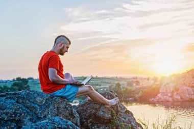Güzel bir gün batımında bir kayanın üzerinde otururken dizüstü bilgisayar üzerinde çalışan bir adam.